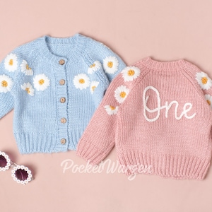 Op de afbeelding: Twee baby-truien, één lichtblauw met witte madeliefjes-borduursels en één roze met witte madeliefjes-borduursels en het woord "Eén" geborduurd op de voorkant.