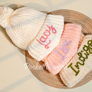 May include: Three knitted hats with pom-poms, stacked on top of each other. The top hat is white with the word "Lady" embroidered in pink. The middle hat is pink with the word "Isa" embroidered in purple. The bottom hat is cream with the word "Indigo" embroidered in green.