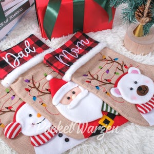 May include: Four Christmas stockings with embroidered designs. The stockings are made of a beige fabric with red and black plaid trim. The stockings are personalised with the names "Mom", "Dad", "Mom", and "No" in white embroidery. Each stocking features a different embroidered design: a snowman, a reindeer, Santa Claus, and a polar bear.