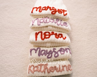 Può includere: Una pila di berretti bianchi lavorati a maglia con nomi ricamati in vari colori. I nomi "Margot", "Natalie", "Nora", "Mayson" e "Katherine" sono visibili. Il testo "Pocket Wansen" è ricamato anche su uno dei berretti.