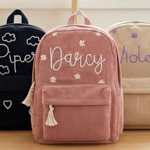 May include: Three corduroy backpacks in navy, pink, and beige. Each backpack has a front pocket and a top handle. The navy backpack has the name "Piper" embroidered in white with cloud and heart details. The pink backpack has the name "Darcy" embroidered in white with flower details. The beige backpack has the name "Violet" embroidered in purple with flower details.