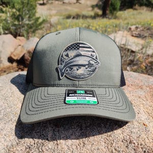 May include: A green and black trucker hat with a patch of a trout jumping out of the water. The patch has a faded American flag design in the background. The hat has a black mesh back and a green front with a black stitched brim. The hat has a label that says "Richardson 112RE Recycled Trucker Fashion".