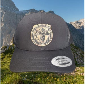 May include: Black mesh trucker hat with a brown and gold embroidered patch featuring a deer in a mountain scene. The hat has a black bill and a black strap with a white "The Authentic Yupoong Snapback Yupoong Since 1974" label.