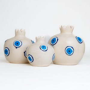 Può includere: Tre vasi in ceramica bianca a forma di melograno con disegni di occhi maligni blu. I vasi sono di dimensioni diverse e sono disposti in fila dal più grande al più piccolo.