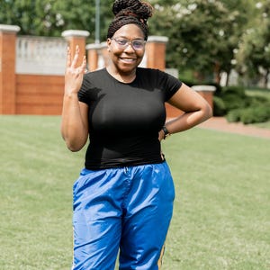 May include: A person wearing a black t-shirt and blue track pants with yellow stripes. The person is smiling and wearing glasses. The background includes a brick building and green grass.