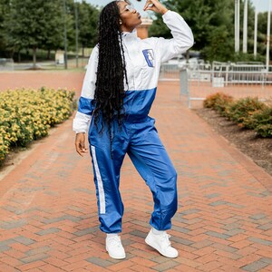May include: A woman wearing a white and blue tracksuit with white stripes down the sides of the legs. She is standing on a brick walkway and looking up at a statue.