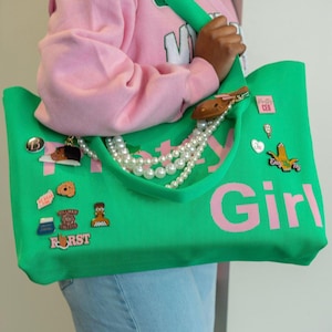 May include: A bright green tote bag with large pink text that reads Pretty Girl. The bag is decorated with various enamel pins, including a brown character, a heart, and a hand gesture, and features a multi-strand white pearl bead accessory.