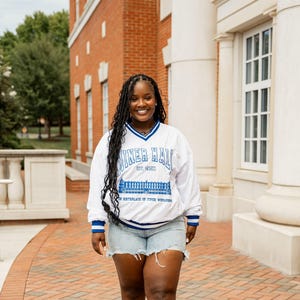 Peut inclure: Un sweat-shirt blanc avec des bordures bleues et le texte "Miner Hall" en bleu. Le sweat-shirt est porté avec un short en jean. L'image est prise à l'extérieur devant un bâtiment en briques.