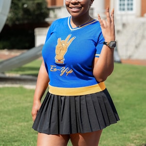 May include: A blue V-neck top with a gold and white trim, featuring a hand gesture graphic and the text "Ee-Yip". The top is paired with a black pleated skirt. The outfit is worn by a person with glasses and a smartwatch.