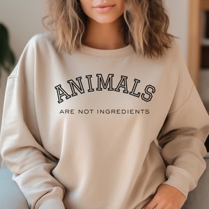 May include: Beige sweatshirt with the text "ANIMALS ARE NOT INGREDIENTS" in black, collegiate-style lettering. The sweatshirt has a crew neck and long sleeves. The overall style is casual and comfortable.