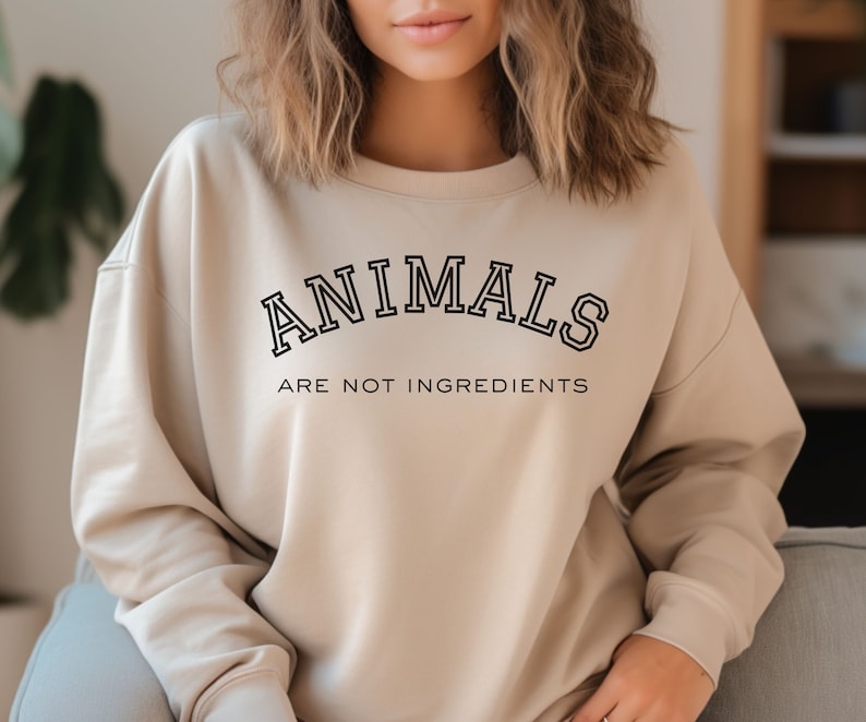 May include: Beige sweatshirt with the text "ANIMALS ARE NOT INGREDIENTS" in black, collegiate-style lettering. The sweatshirt has a crew neck and long sleeves. The overall style is casual and comfortable.
