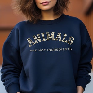 May include: Navy blue sweatshirt with the text "ANIMALS ARE NOT INGREDIENTS" in a cream-colored, collegiate-style font. The sweatshirt has a crew neck and long sleeves, and is made of a soft material.