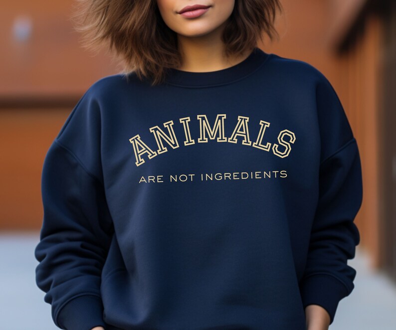 May include: Navy blue sweatshirt with the text "ANIMALS ARE NOT INGREDIENTS" in a cream-colored, collegiate-style font. The sweatshirt has a crew neck and long sleeves, and is made of a soft material.