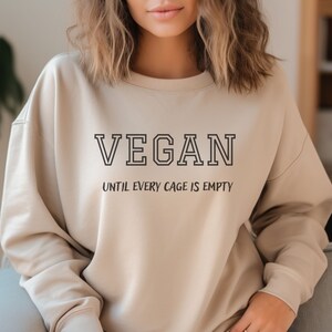 May include: A beige sweatshirt with the text "VEGAN UNTIL EVERY CAGE IS EMPTY" printed on the front.
