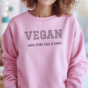 May include: Pink sweatshirt with the text "VEGAN UNTIL EVERY CAGE IS EMPTY" printed on the front.