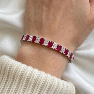 Könnte beinhalten: Ein Tennisarmband mit abwechselnd rechteckig geschliffenen roten Edelsteinen und klaren, rechteckig geschliffenen Steinen in einem Metallband. Das Armband wird am Handgelenk getragen und zeigt ein wiederkehrendes Muster aus leuchtend roten und klaren Steinen.