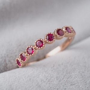 May include: A rose gold band ring featuring a row of seven round-cut red rubies. Each ruby is individually set within a milgrain-detailed bezel setting, creating a continuous line of gemstones along the top half of the polished metal band.