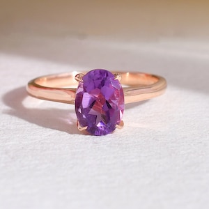 May include: A rose gold ring featuring an oval-cut, deep purple amethyst gemstone. The ring's band is a delicate, polished metal, and the gemstone is the focal point, set in a classic four-prong setting. The ring is displayed on a white surface.