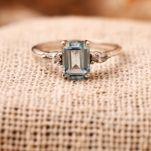 May include: A silver-toned ring featuring a central emerald-cut light blue gemstone flanked by two smaller, clear pear-shaped stones on each side, set on a thin, polished metal band that provides a clean and structured appearance.