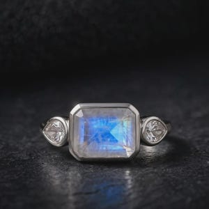 May include: A silver-toned ring featuring a central rectangular moonstone with a blue iridescent sheen, flanked by two smaller pear-shaped clear stones. The stones are set in a smooth metal bezel, creating a clean and polished appearance.