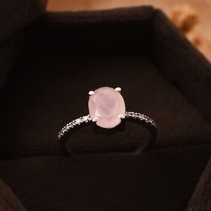 Könnte beinhalten: Ein zarter Ring mit einem blassrosa ovalen Edelstein, der von einer Reihe kleinerer, klarer Edelsteine umgeben ist. Der Ring wird in einer braunen Samt-Ringbox präsentiert.
