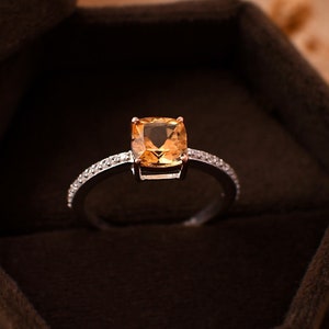 Puede incluir: Un anillo de oro con una gran piedra de topacio de corte cuadrado engastada en una banda de metal blanco. El anillo está rodeado de diamantes más pequeños y se presenta en una caja de anillo de terciopelo marrón.