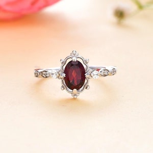 May include: A silver ring with a red oval gemstone surrounded by smaller white gemstones. The ring has a delicate, ornate design.