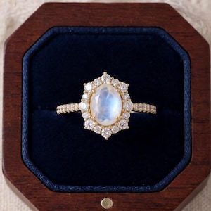 May include: A gold ring featuring a central oval-shaped moonstone cabochon surrounded by a halo of small round brilliant-cut diamonds. The band is thin and also set with small diamonds, presented inside a dark brown wooden octagonal box.