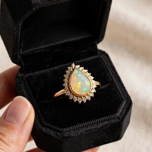 May include: A gold ring featuring a pear-shaped opal center stone surrounded by a halo of small round white diamonds. The ring is presented inside a black velvet octagonal jewelry box with a hinged lid and a soft black interior lining.