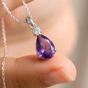 Puede incluir: Un collar de tono plateado con un colgante de amatista morada tallada en forma de pera, suspendido de una cadena fina. Una pequeña piedra transparente de corte redondo está engastada sobre la amatista, uniendo el colgante a la cadena.