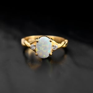 Op de afbeelding: Een gouden ring met een witte opaal-edelsteen en twee kleinere heldere edelstenen. De ring heeft een gedraaide band.