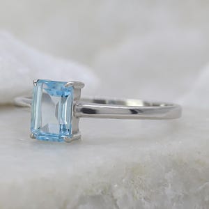 Op de afbeelding: Een zilveren ring met een rechthoekige, lichtblauwe edelsteen. De edelsteen is het middelpunt, gezet in een eenvoudige, elegante band. De ring wordt getoond op een wit, getextureerd oppervlak, dat de kleur en het ontwerp benadrukt.