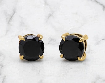 May include: A pair of stud earrings featuring round, faceted black stones set in four-prong gold metal settings. The earrings have a polished finish and a classic circular design, showcasing the contrast between the dark stones and gold.
