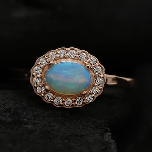 May include: A rose gold ring featuring a large oval opal gemstone set within a halo of smaller diamonds.