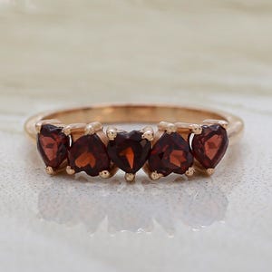 Könnte beinhalten: Ein roségoldener Ring mit fünf herzförmigen, tiefroten Edelsteinen. Die Edelsteine sind in einer Reihe auf der Oberseite des Rings angeordnet. Der Ring ist vor einem weichen, neutralen Hintergrund zu sehen.