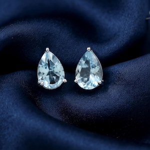 May include: A pair of stud earrings featuring faceted, pear-shaped light blue gemstones set in polished silver-toned metal. Each earring has a pointed top and a rounded base, showcasing clear, reflective surfaces that catch the light.