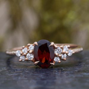Op de afbeelding: Roségouden ring met een ovale, dieprode edelsteen in het midden. De ring is versierd met kleine, ronde, heldere edelstenen die aan weerszijden van de hoofdsteen zijn gegroepeerd. De ring is geplaatst tegen een vage groene en grijze achtergrond.