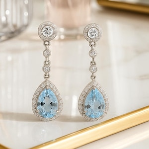 May include: A pair of drop earrings with light blue, pear-shaped gemstones, each encircled by small, clear stones. The earrings are linked by small, round, clear stones and a larger, round clear stone at the top. The earrings are set on a gold-edged tray.