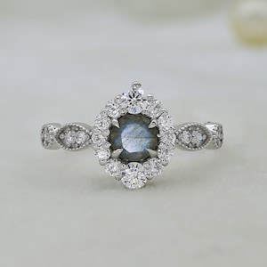 May include: A silver ring with a large, oval-shaped, dark blue labradorite gemstone surrounded by a halo of smaller, round, clear gemstones. The band is decorated with a delicate, floral design.