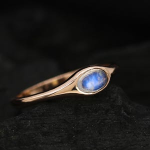 May include: A rose gold ring featuring a blue moonstone in an oval setting. The ring's band curves gracefully, and the moonstone has a soft, milky appearance. The ring is set against a dark, textured background.