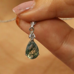 May include: A silver necklace with a teardrop-shaped pendant featuring a green and white moss agate stone. The stone has a unique pattern of green and white swirls. The pendant is accented with a small clear crystal.
