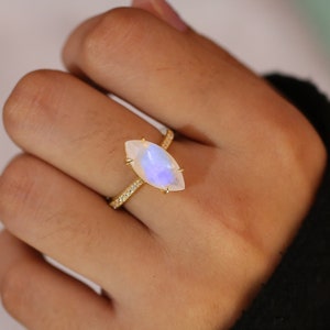 Op de afbeelding: Een gouden ring met een in marquise geslepen maansteen in het midden met een blauwe iriserende glans. De dunne ringband is bezet met een rij kleine, heldere pavé-steentjes langs de schacht, vastgezet door vier fijne pootjes op de steen.