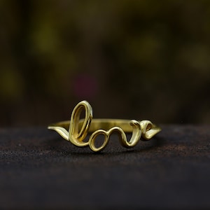 May include: A gold-colored ring with the word "love" in a cursive script. The ring is made of a shiny, reflective metal and sits on a dark surface. The word "love" is the primary design element.