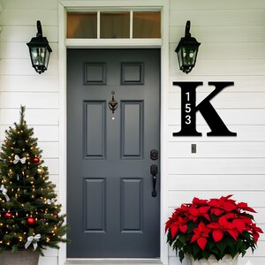 May include: Black metal house number sign featuring the letter "K" with the numbers "153" in white. The sign is mounted on a white wall next to a dark gray front door. A Christmas tree and red poinsettias decorate the porch.