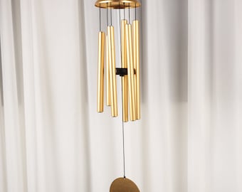 Könnte beinhalten: Ein goldfarbener Windspiel mit einem kreisförmigen Oberteil und einer Reihe von zylindrischen Röhren, die darunter hängen. Eine runde, flache Scheibe hängt unten. Die Klangspiele sind an schwarzen Schnüren vor einem weißen Vorhanghintergrund aufgehängt.
