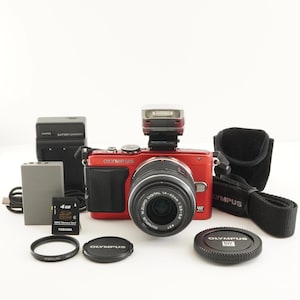 May include: A red Olympus digital camera with a mounted flash, a 14-42mm lens, a black battery charger, a grey battery, a 4GB Toshiba SD card, a black camera strap, a lens filter, and two black Olympus lens caps.