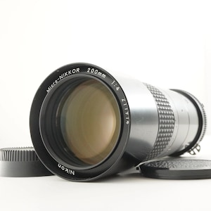 May include: A Nikon Micro-NIKKOR 200mm f/4 camera lens. The lens is silver and black with a large front element. The lens cap is on the left, and the lens is labeled with the Nikon logo and other specifications.