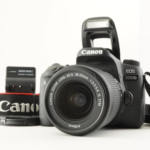 Op de afbeelding: Een zwarte Canon EOS 9000D digitale camera met een EF-S 18-55mm 1:3.5-5.6 IS STM zoomlens en een uitgeklapte flitser. Inbegrepen zijn een zwarte camerariem met het Canon-logo, een lensdop en een zwarte Sixoctave-batterijlader.