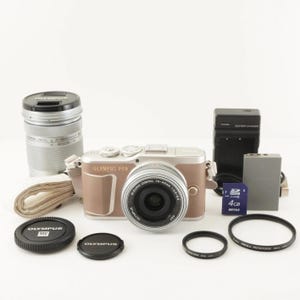 May include: An Olympus PEN E-PL7 camera kit with a brown and silver body, lens, and accessories. Includes a lens cap, battery charger, SD card, and lens filters. The camera has a 14-42mm lens.