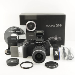 Może przedstawiać: Czarny zestaw aparatu Olympus OM-D z różnymi akcesoriami, w tym obiektywem, lampą błyskową, ładowarką i płytą CD-ROM. Zestaw zawiera pasek do aparatu z napisem "OM-D" oraz czarne pudełko z napisem "OLYMPUS OM-D".